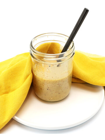 Small glass jar of honey mustard sauce sitting on a plate with a spoon resting in the jar and a yellow towel laying next to the jar.