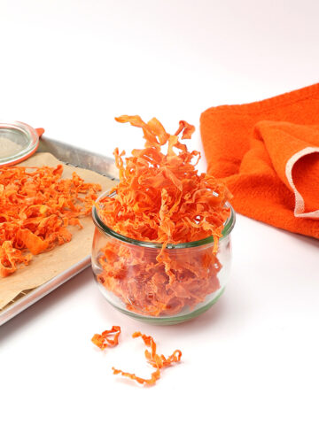 light and airy carrot chips a.k.a. carrot crisps in a small bowl and some on a baking sheet with an orange towel next to them.