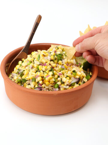 Corn salsa in a terra cotta clay pot with a wooden spoon and a hand holding a tortilla chip.