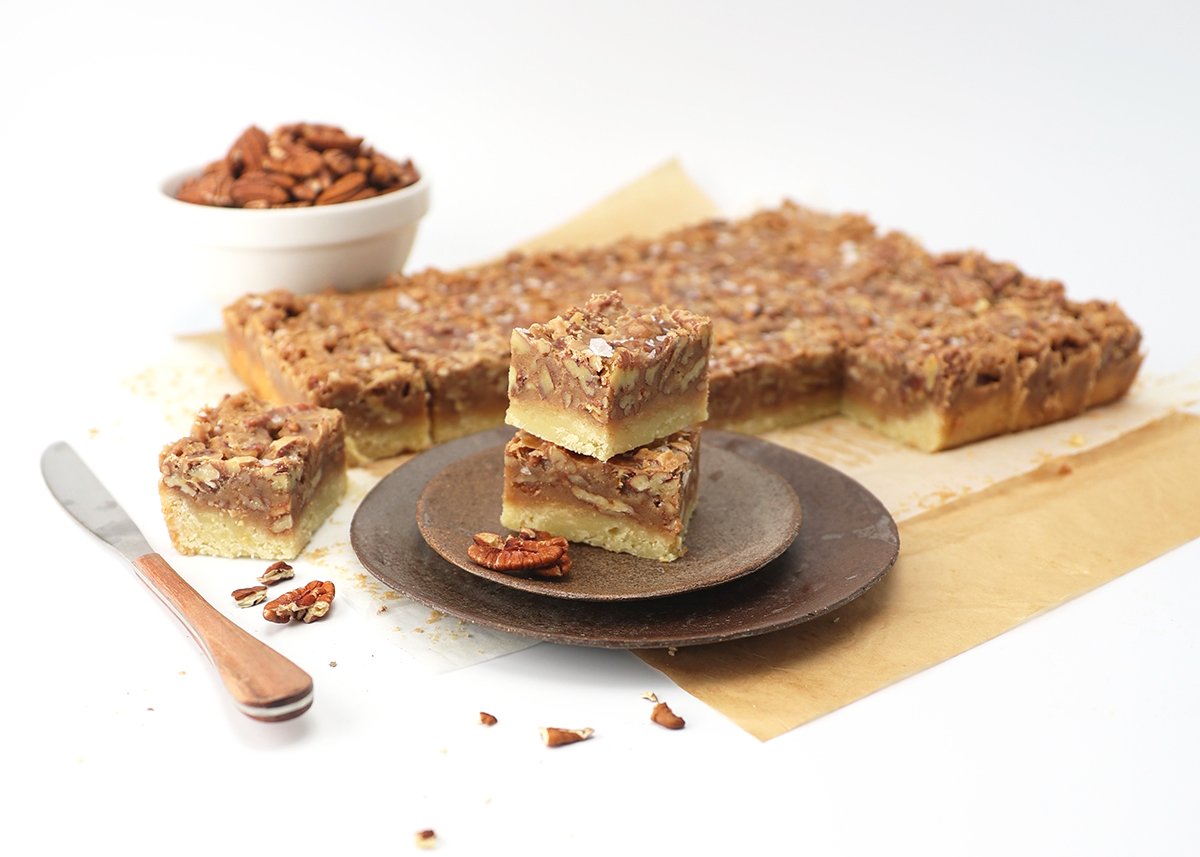 Two square pieces of pecan pie bars stacked on top of one another on a plate with the remaining bars laying next to it on parchment paper.