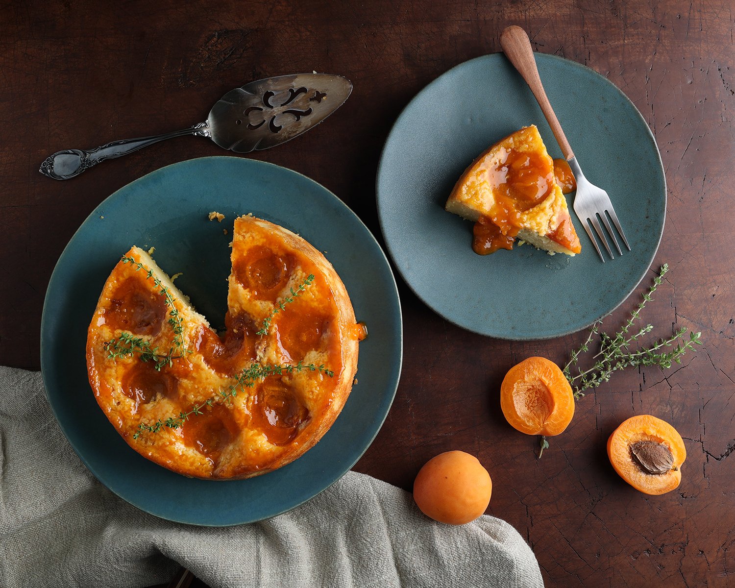 A slice of Apricot ricotta thyme cake is served on a small plate next to the freshly baked summer cake and halved apricots laying next to the plate.