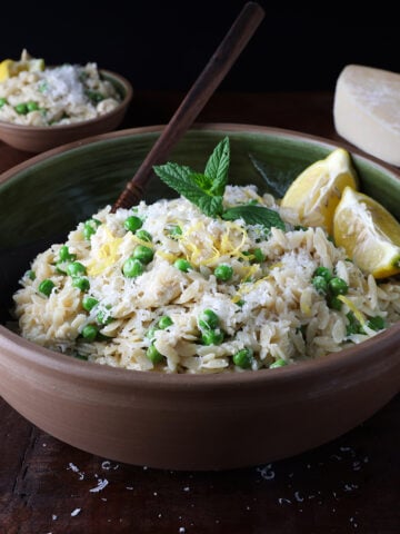 Fresh parmesan grated over a large wooden bowl of lemon ricotta orzo with baby peas.