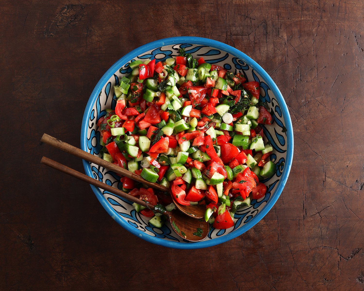 Salad serving spoons resting in a bowl of fresh Israeli cucumber tomato salad.