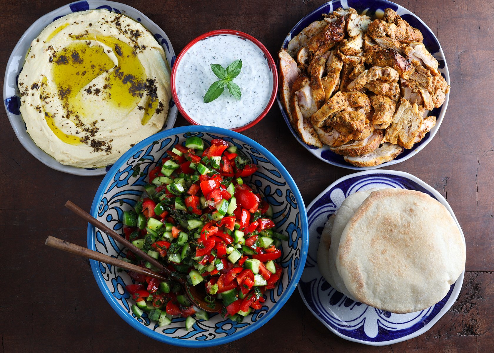 Naan bread, slices of seasoned chicken breast, fresh hummus and an Israeli tomato and cucumber salad all in serving dishes.