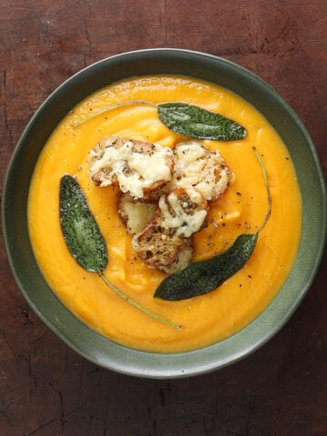Close up of a bowl of butternut squash soup with small cheesy croutons placed in the middle and crispy sage leaves around them.
