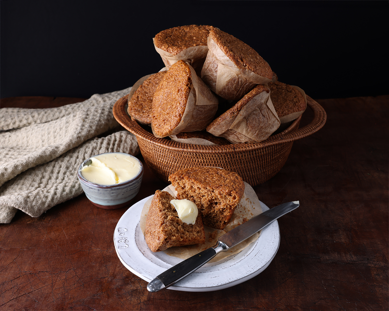 A small plate holding an apple bran muffin split in half with a pat of butter on it next to the basket of freshly baked muffins.