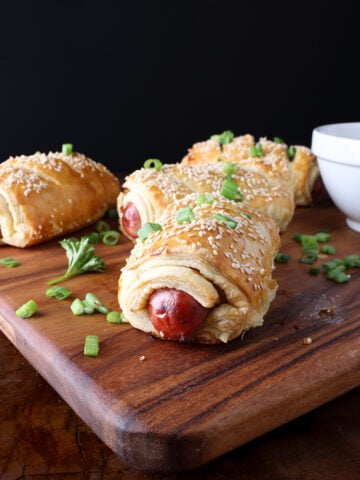 Golden brown baked pigs in a blanket with caramelized onions and topped with sesame seeds and green onions.