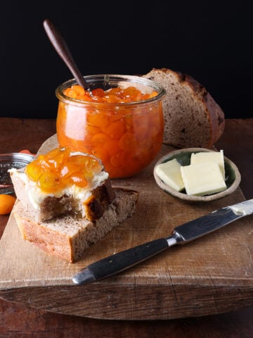 Two thick slices of homemade bread next to a small plate of butter and a glass jar of kumquat jam.