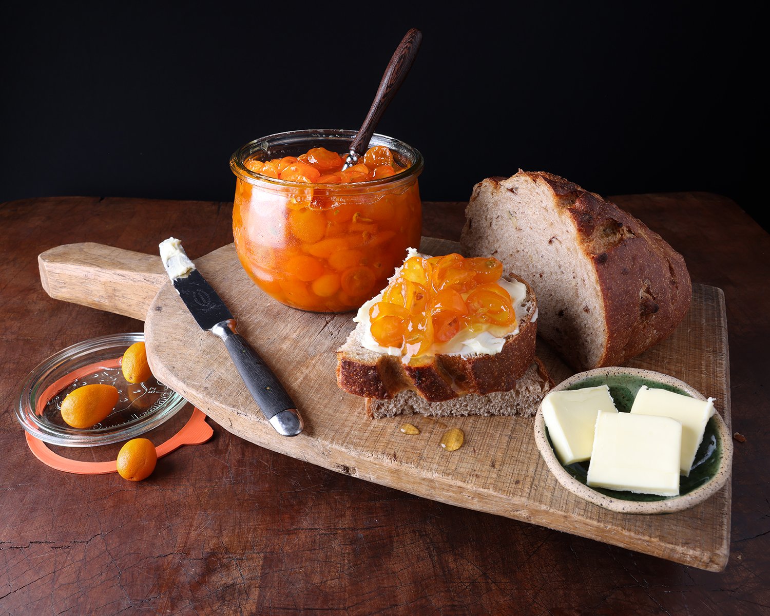 Half a loaf of homemade bread next to a slice of buttered bread topped with a topping of rich kumquat jam.