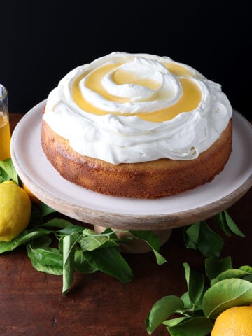 Lemon syrup in a glass pitcher next to a lemon cake topped with whipped cream and lemon syrup drizzled over.