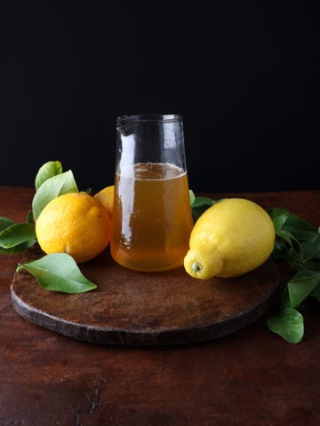 Bright lemons and green leaves garnished around a glass pitcher of lemon syrup.