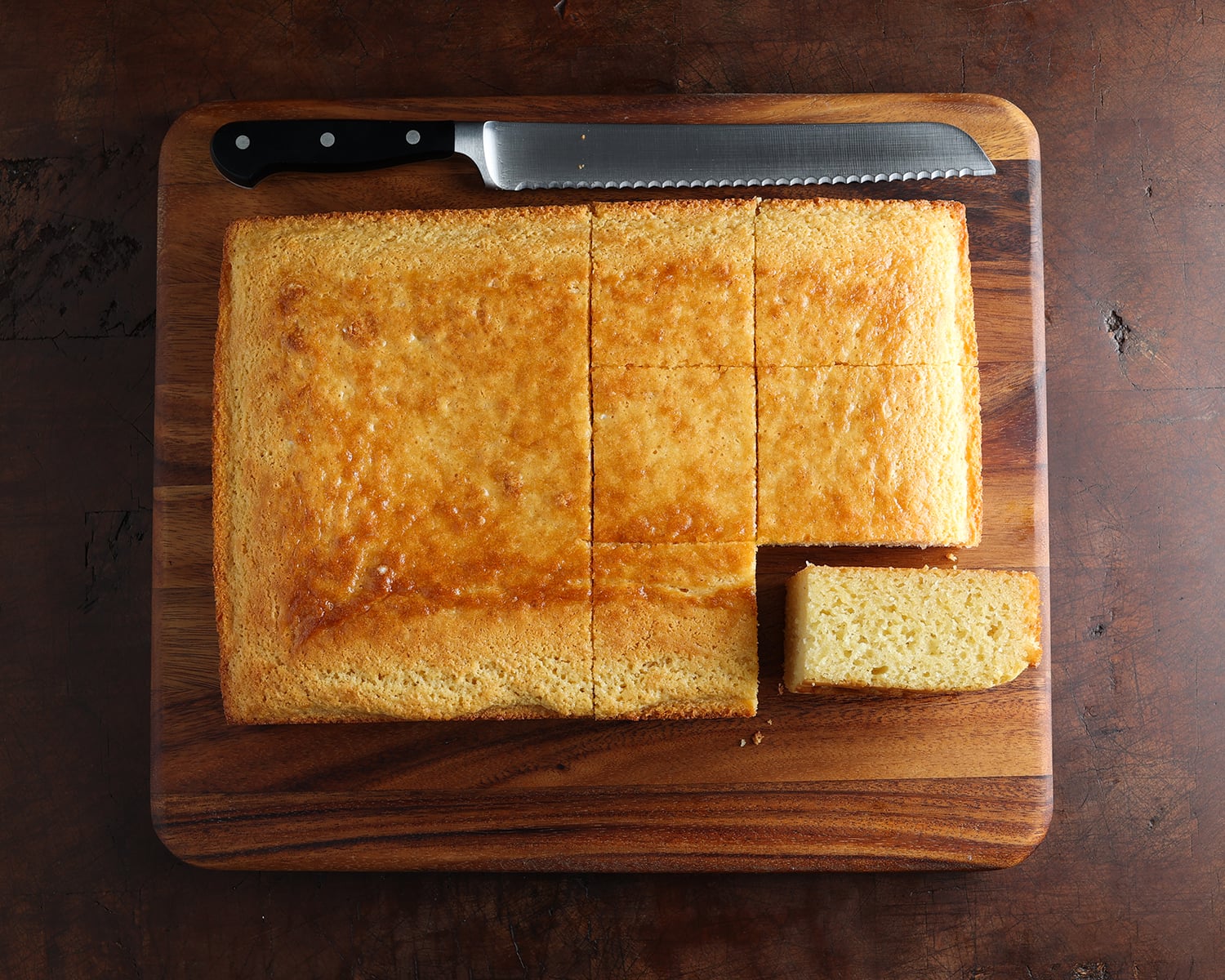 Cornbread resting on a wooden cutting board, with cut marks to serve square pieces and a bread knife laying on its side.
