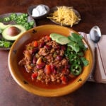 Bowl of smoky chipotle chili garnished with avocado slices, jalapeno rounds and cilantro leaves.