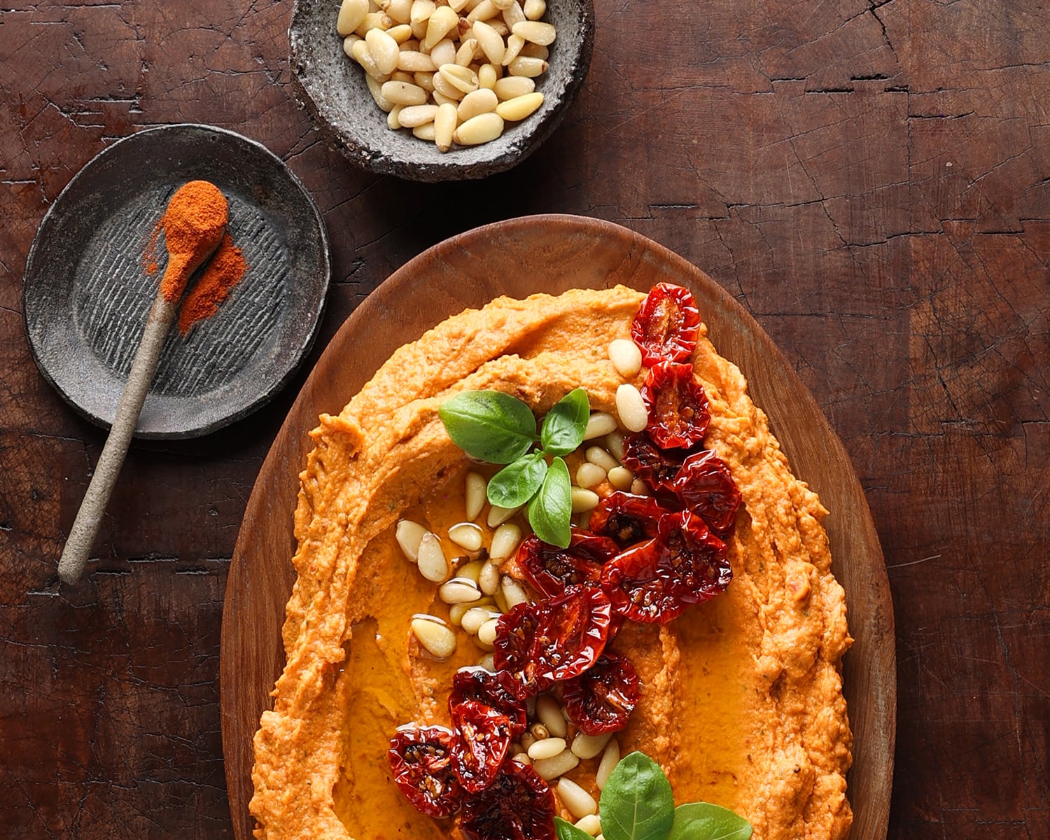 Partial view of a wooden bowl with a homemade sun-dried tomato hummus with a garnish of pine nuts, fresh basil leaves and sun-dried tomatoes.