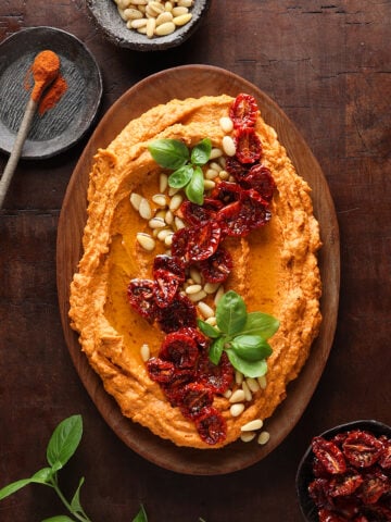 Hummus prepared with sun-dried tomatoes, pine nuts and fresh basil shared on a wooden bowl.