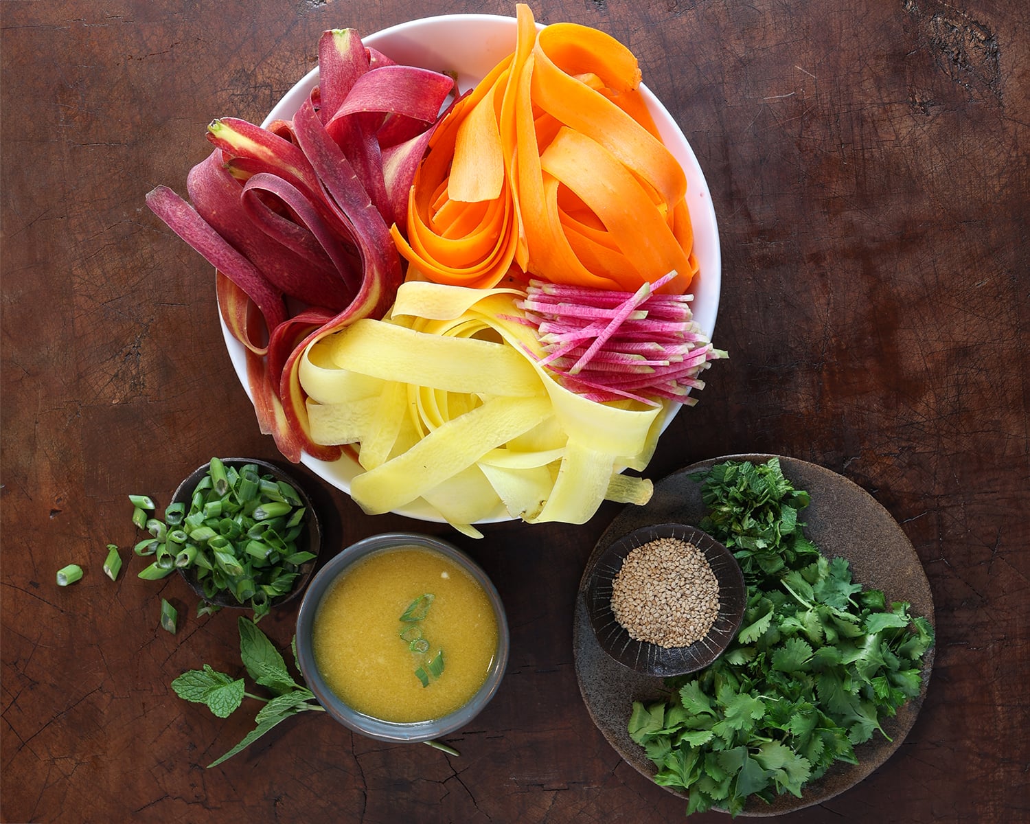 Ribboned carrots in a bowl, chopped fresh ingredients on a plate and homemade dressing with a small bowl of chopped green onions.