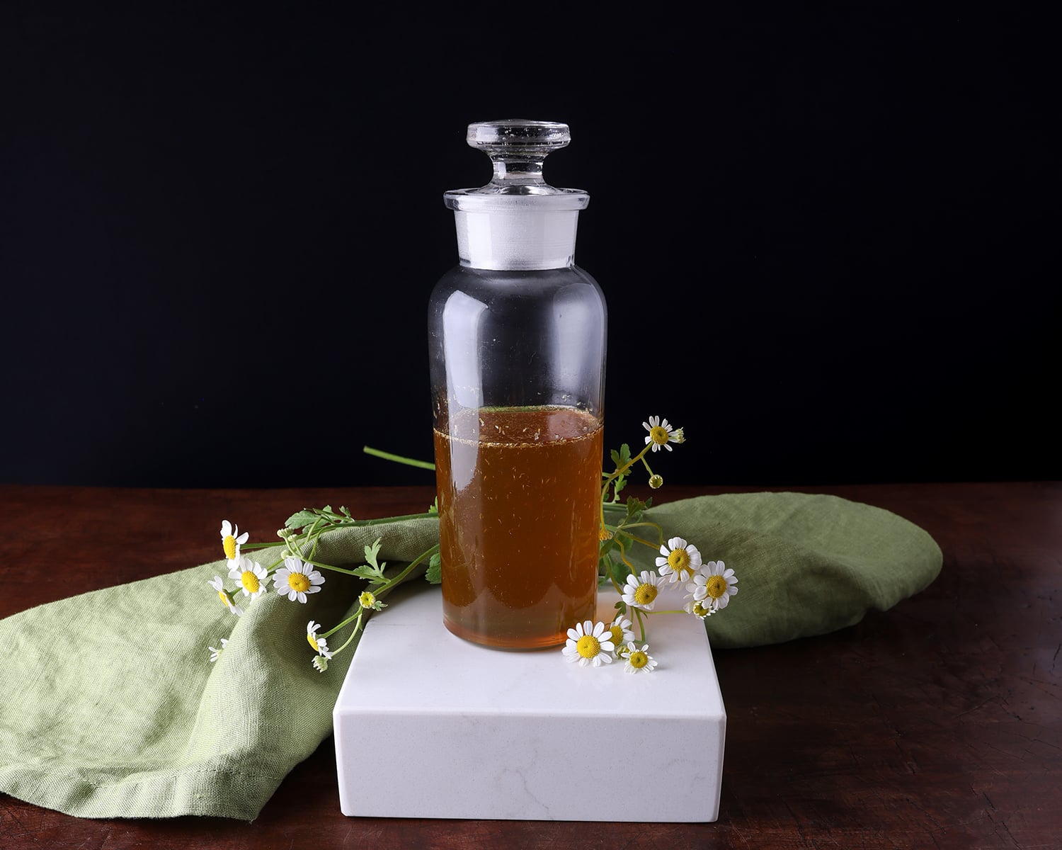 Chamomile syrup in a glass container with small white flowers laying around it.