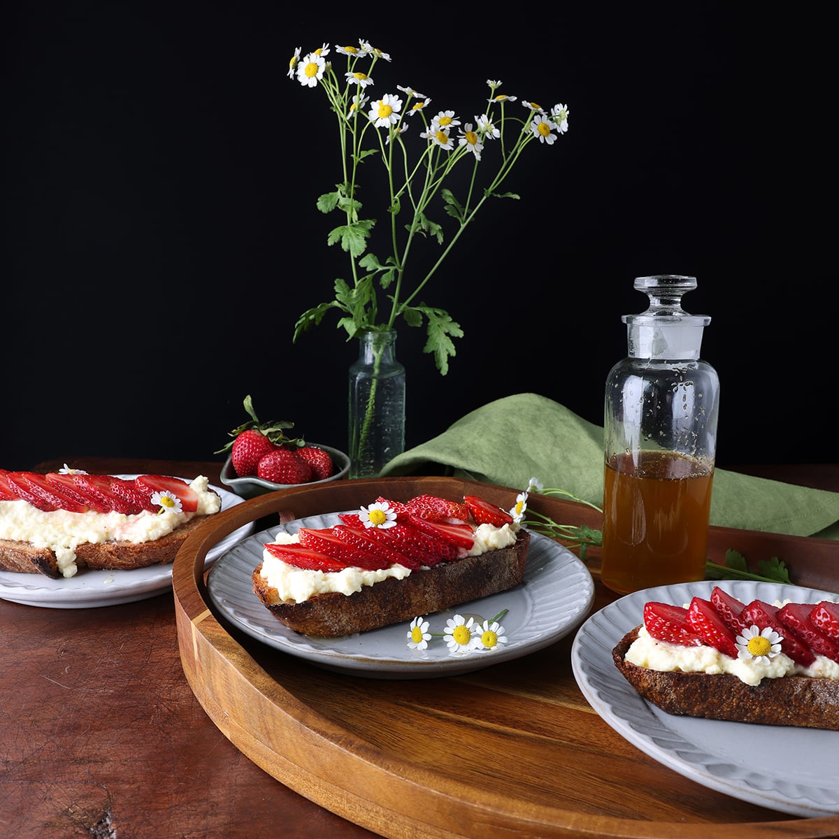 Side view of a wooden serving tray holding strawberry ricotta toast and a small vase of flowers next to it.