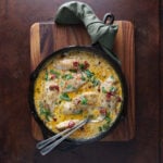 Cast iron skillet with a couple of spoons resting in a creamy sauce with chicken breasts resting in the mixture.