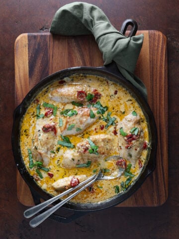 Cast iron skillet with a couple of spoons resting in a creamy sauce with chicken breasts resting in the mixture.