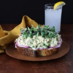 Thick slice of bread topped with slices of red onions, smashed chickpea salad and garnished with microgreens.