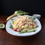 Roasted corn on the cob laying next to a large plate of grilled corn pasta salad with lime wedges garnished on the side.