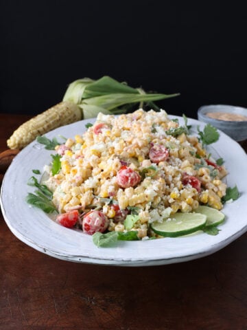 Roasted corn on the cob laying next to a large plate of grilled corn pasta salad with lime wedges garnished on the side.