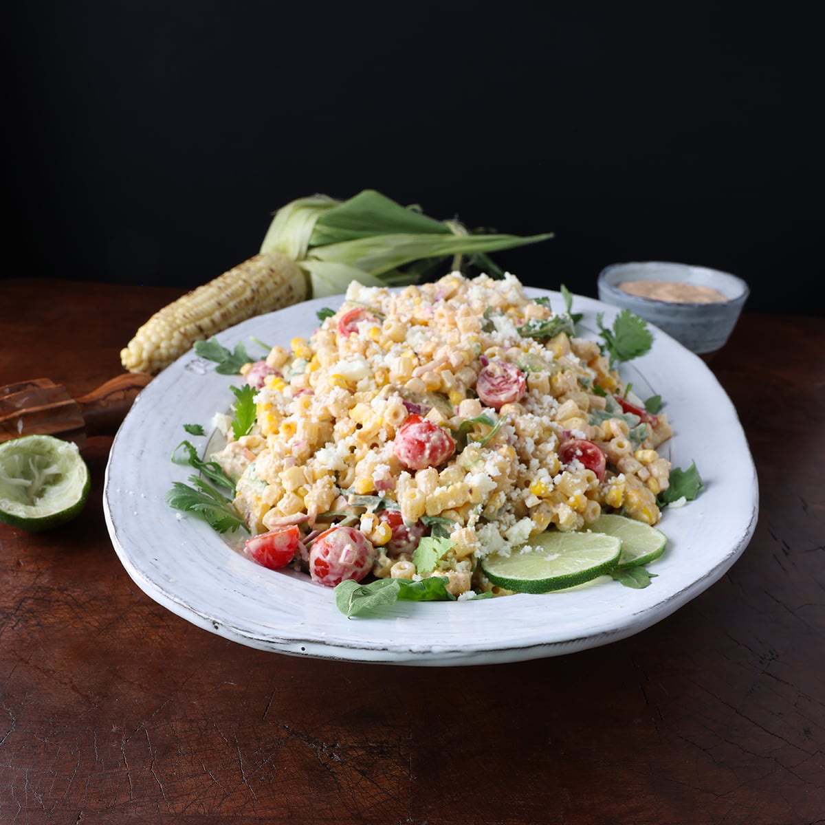 Roasted corn on the cob laying next to a large plate of grilled corn pasta salad with lime wedges garnished on the side.