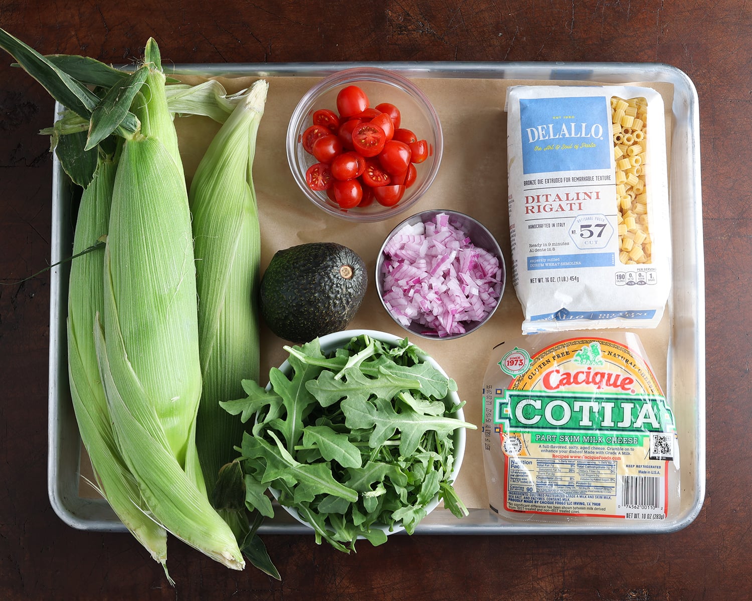Whole ingredients used to prepare a grilled corn salad are laid out on a baking sheet.
