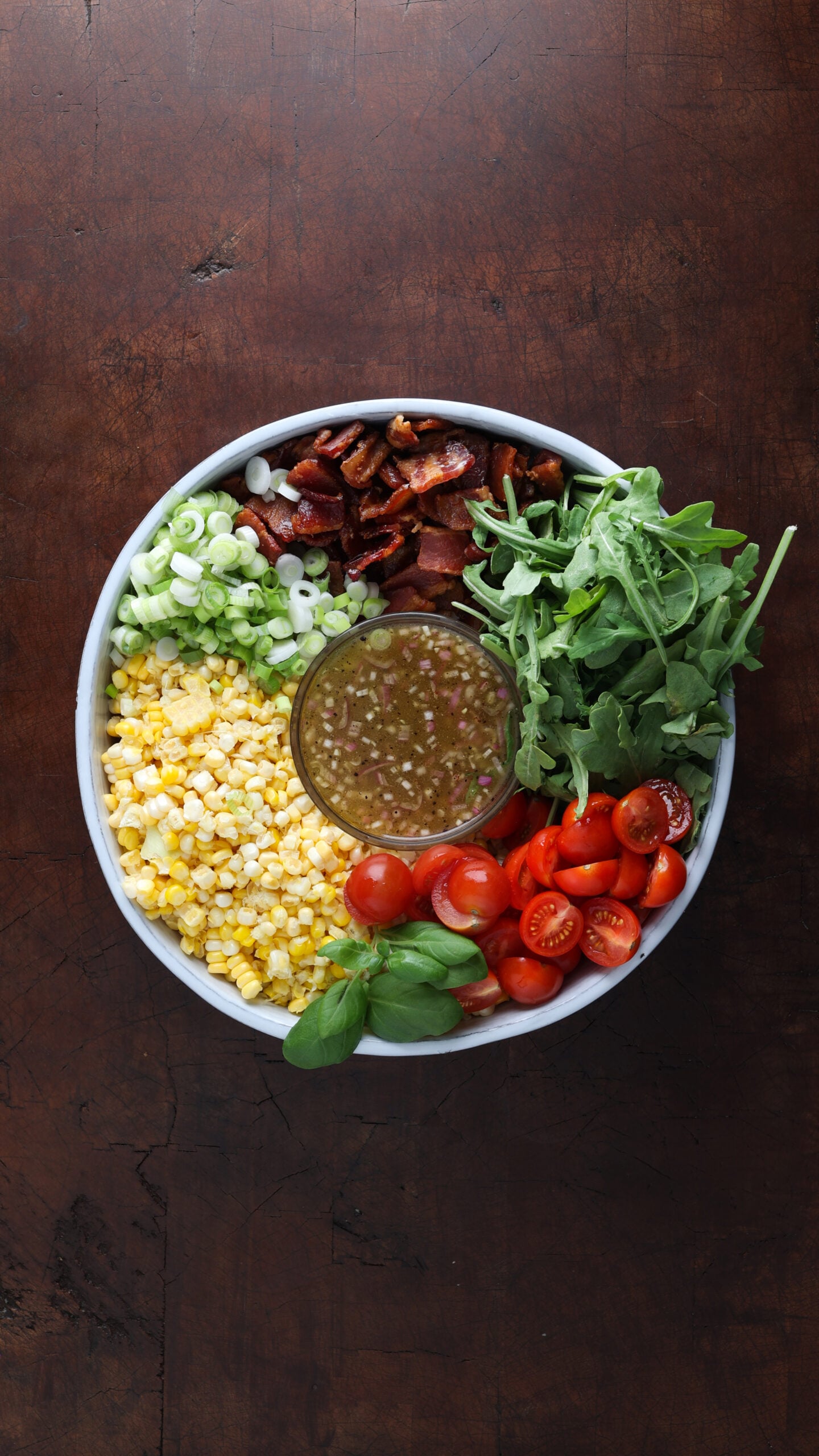 Top view of a round platter with the individual ingredients chopped, sliced and put in the container but not mixed for a BLT corn salad and the dressing in the middle in a bowl.
