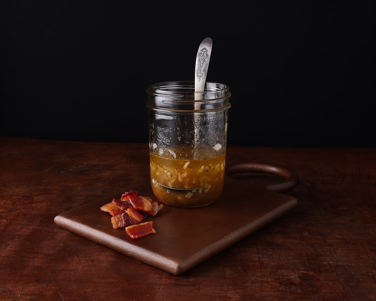 Chopped crispy bacon pices resting next to a pint glass jar partially filled with bacon shallot vinaigrette and a spoon resting in the mixture.