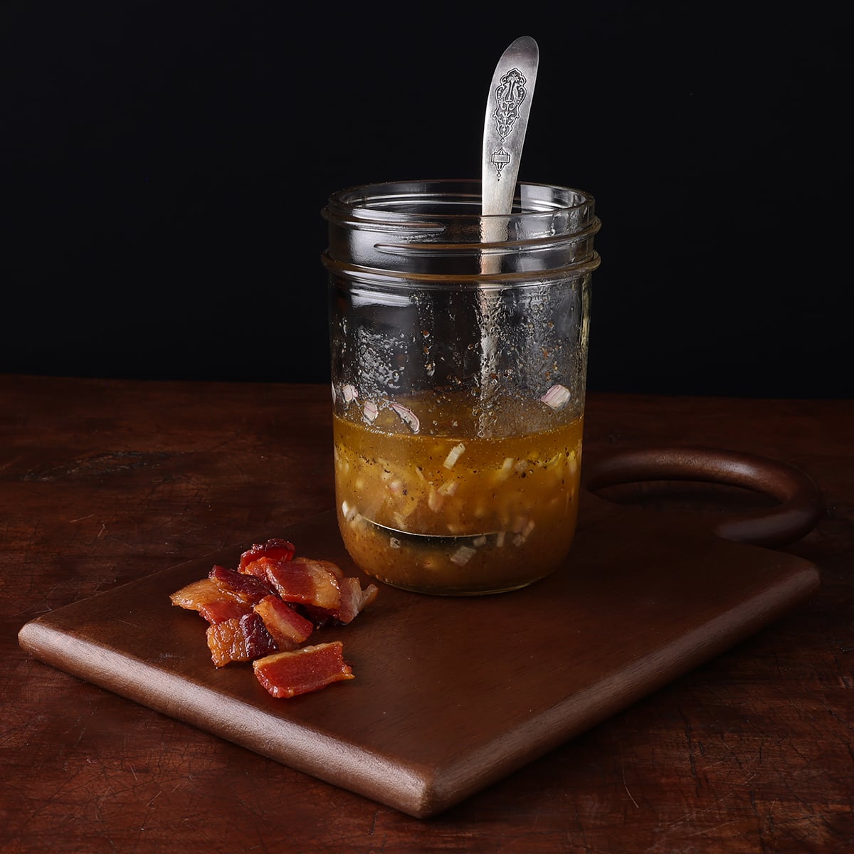 Warm Bacon Sallot Vinaigrette in a mason jar on a wood cutting board with cooked bacon pieces