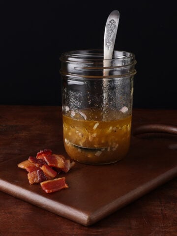 Wooden serving board holding a partially filled glass pint jar with bacon shallot vinaigrette.