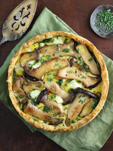 A freshly baked potato crusted quiche with king trumpet mushrooms and goat heese on a sage green linen with a silver serving dish resting on the side.