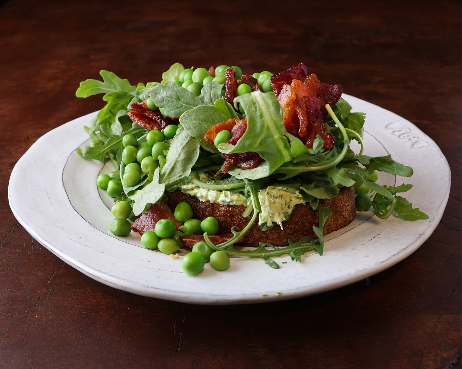 Creamy herbed goat cheese on a thick slice of bread with arugula, crispy bacon pieces and English peas on top and spilling onto the side of the toast.