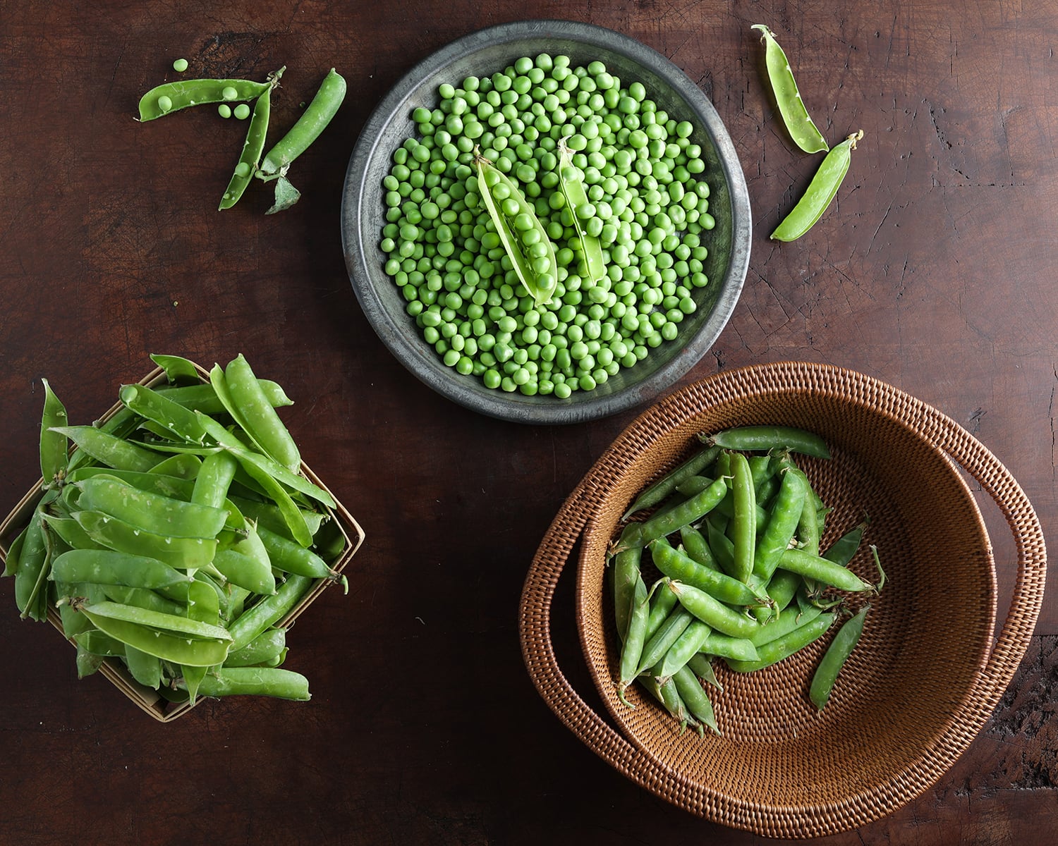 English peas in their pods in one bowl, hte peas removed and shelled into a bowl and the empty pea pods in a basket.