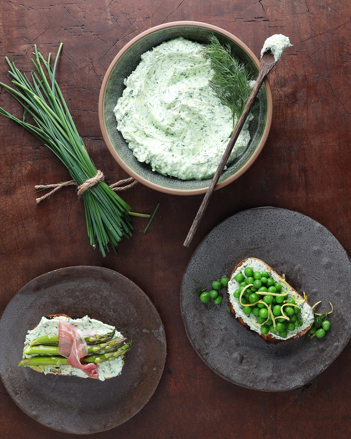 Herbed goat cheese in a round dish with some smeared over a piece of toast with English peas and another topping toast with asparagus and pancetta