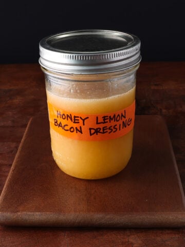 Closed lid on a pint glass jar with tape that says Honey Lemon Bacon Dressing and shows an orangey liquid inside.