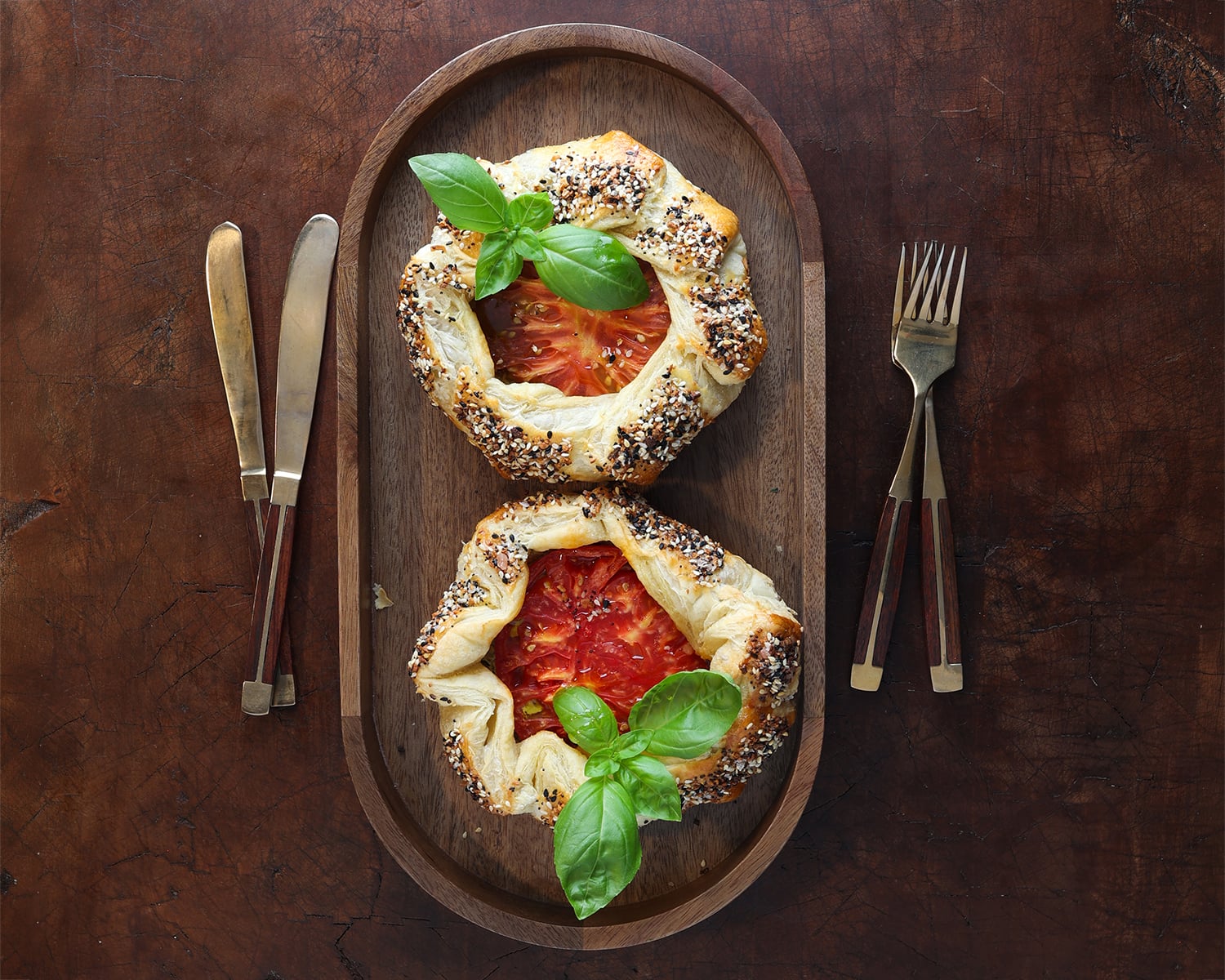 Fork and knife resting besides a wooden serving tray with two puff pastry tomato galettes.