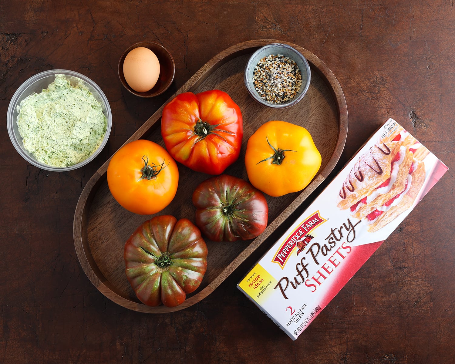 Fresh heirloom tomatoes, everything bagel seasoning, an egg, puff pastry sheets and herby goat cheese spread are laid out to show what's needed to prepare tomato galettes.