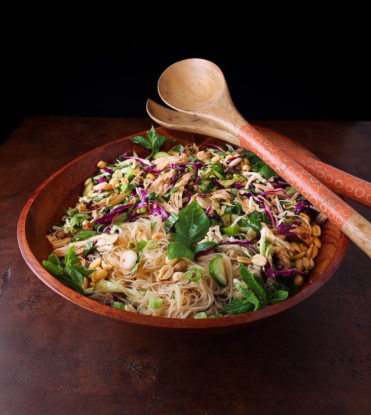 Wooden salad tongs resting on the edge of a wooden salad bowls that's serving an herb chicken vermicelli salad with a vietnamese inspired dish.