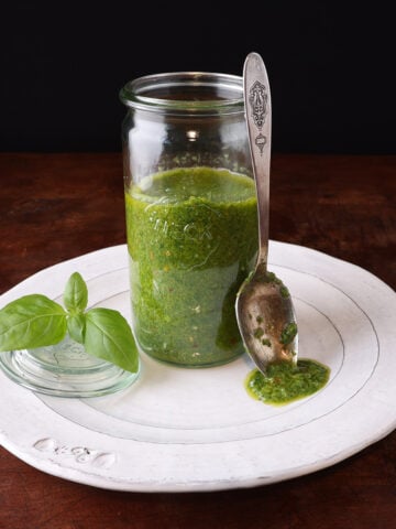 Glass jar with a homemade basil vinaigrette and a spoon with the sauce laying on the edge of the glass, with the sauce dripping onto a white plate.