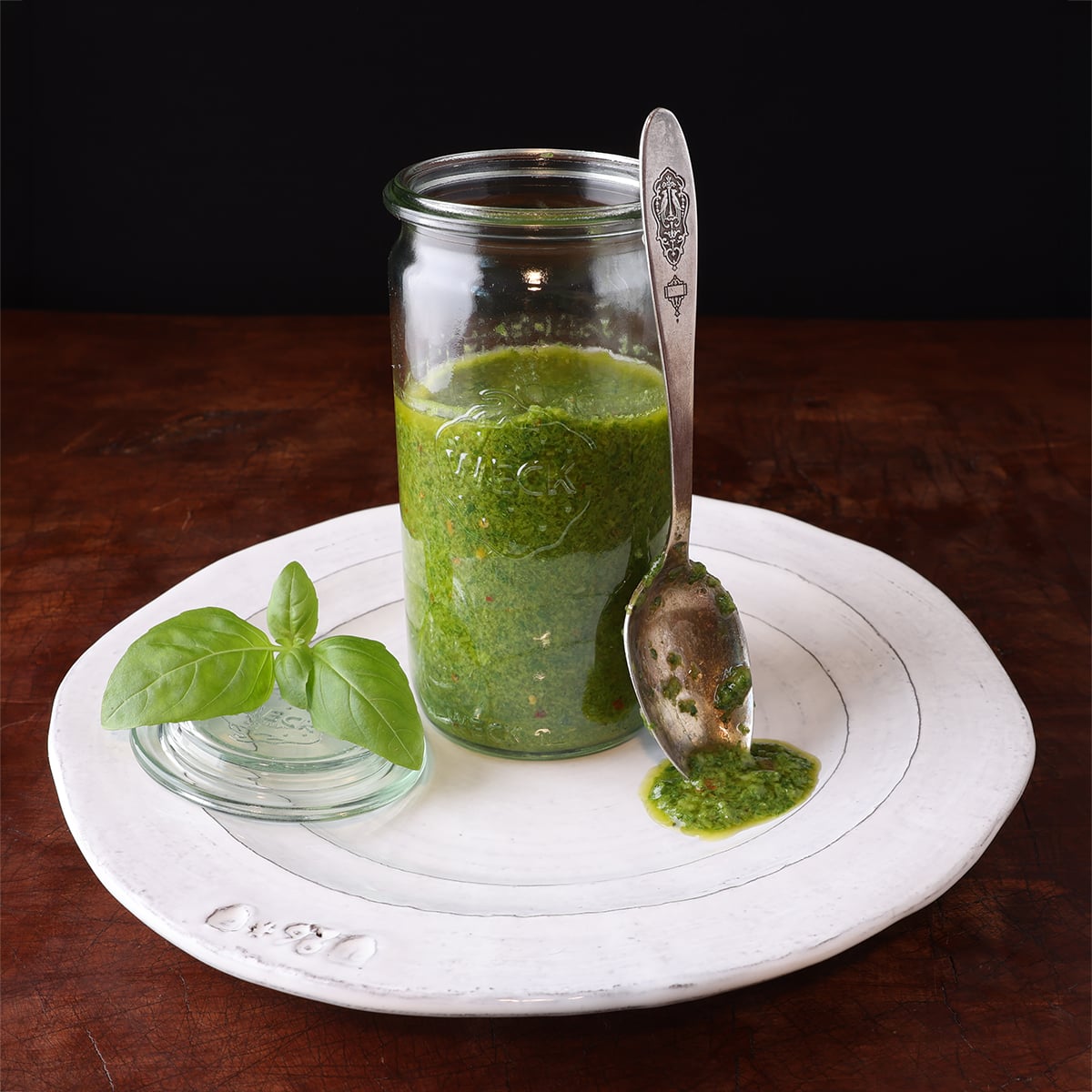 Glass jar with a homemade basil vinaigrette and a spoon with the sauce laying on the edge of the glass, with the sauce dripping onto a white plate.