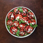 Thin sliced tomatoes laid out on a plate with feta cheese, pine nuts and balsamic glaze over top.