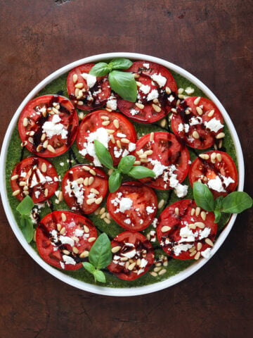 Thin sliced tomatoes laid out on a plate with feta cheese, pine nuts and balsamic glaze over top.