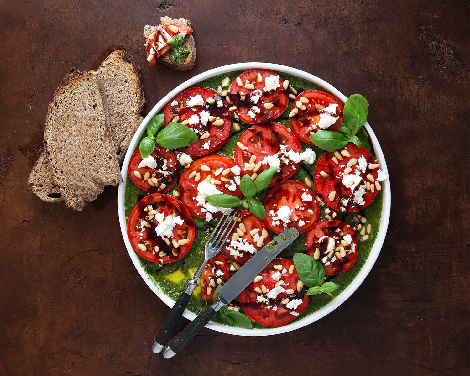 Thin sliced tomato Carpaccio laid out on a plate with basil vinaigrette drizzled on top and fresh basil sprigs laid over the top in a few spots.