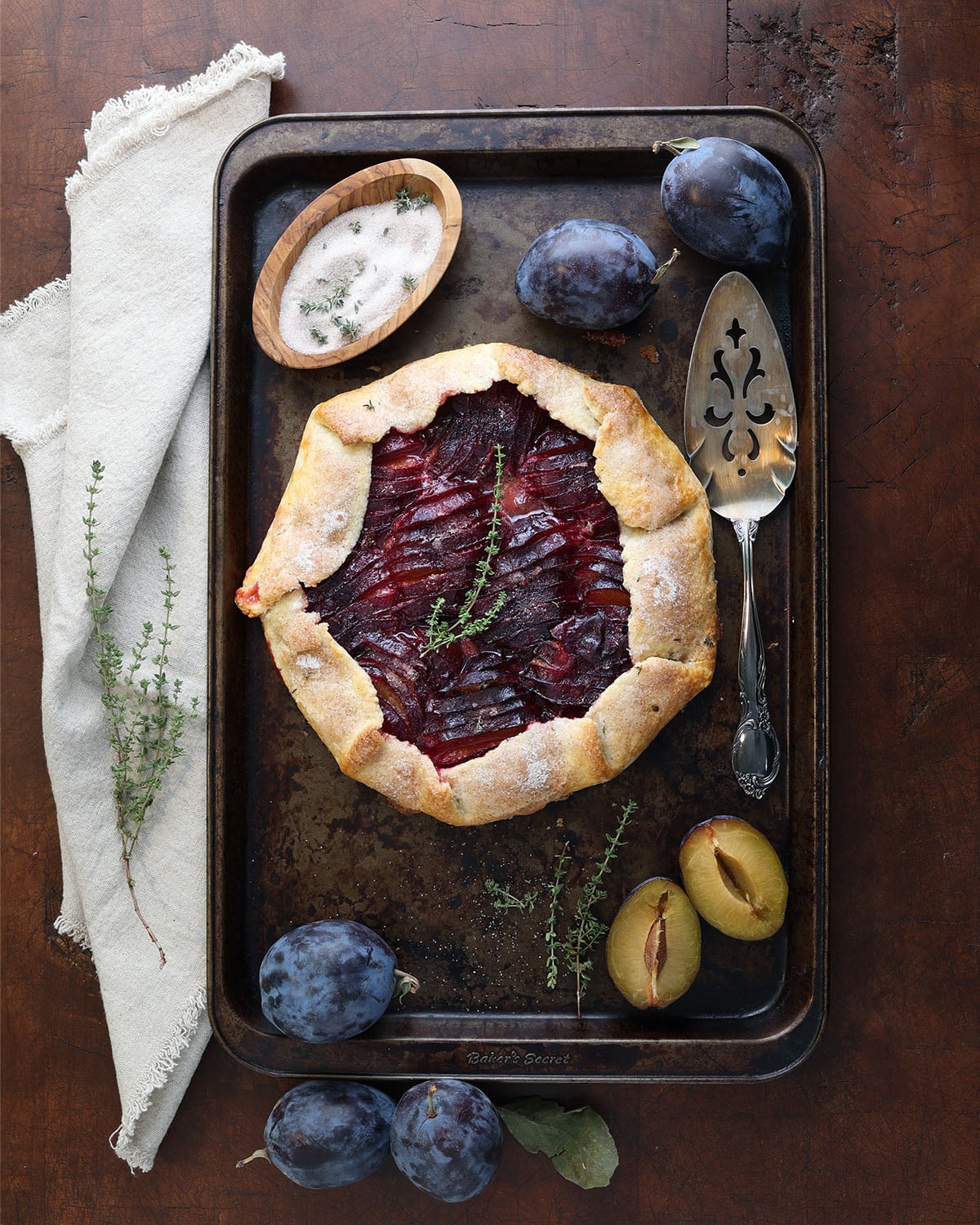 Empress plum galette on rustic pan with a pie server, fresh scattered plums and fresh thyme and small bowl of spiced thyme sugar