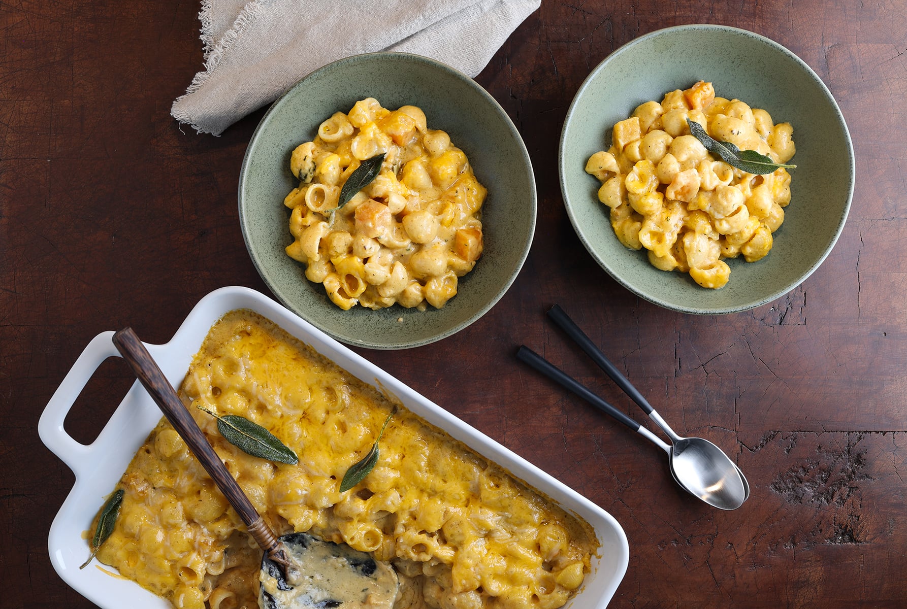 Baking dish of butternut squash mac and cheese with a serving spoon resting in it and two bowls of the pasta served alongside.