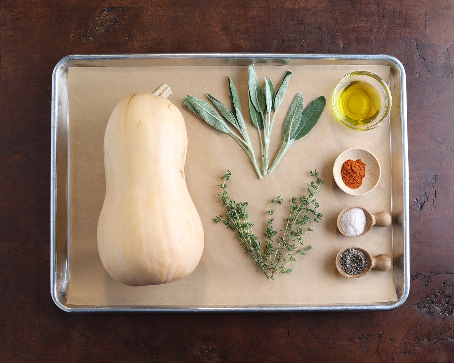 Fresh herbs, a butternut squash, and spices measured out on a baking tray to show.