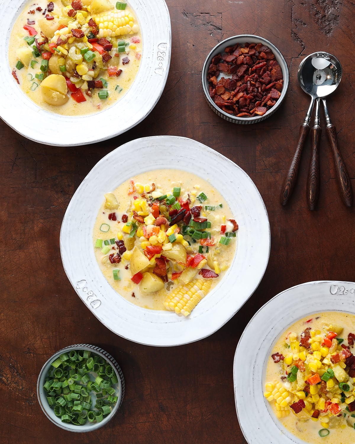 Spoons resting next to some bowls of Fresh corn chowder soup with crispy bacon pieces and green onions garnished over top.
